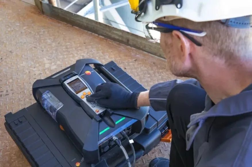 A technician in gloves operates a handheld device connected to a base unit on a textured metal surface in an industrial setting.