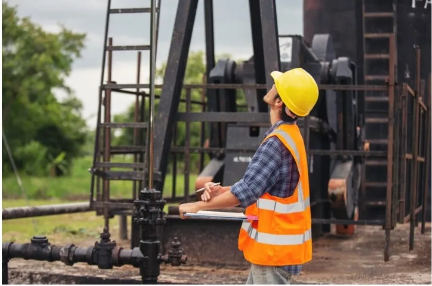 A worker in a yellow hard hat and safety vest observes equipment at a construction site, holding a clipboard and noting details.
