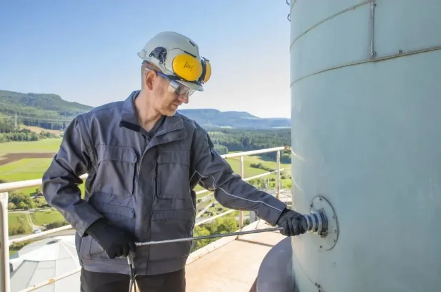A worker in safety gear operates machinery atop a tall structure, with a scenic landscape of fields and hills in the background.